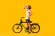 Young male cyclist with bicycle drinking water on yellow background