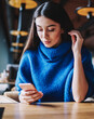 Concentrated teenage hipster girl in blue sweater using mobile phone for blogging indoors
