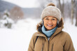 © pangamedia - A senior latin woman is posing in front of the camera happily with a winter coat and a winter hat in a in snow covered country landscape during day in winter while snowing