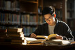 © pangamedia - A senior male asian student is studying while wearing glasses with a book in a busy school library on a table with a bookcase in the background