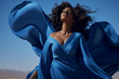 © pangamedia - A beautiful stunning african american woman is wearing a dress with eyes closed with flying waving blue fabric with a desert as background : a full waving dress