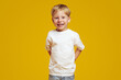 Positive blonde boy in casual white t-shirt keeping hands behind back and smiling for camera while standing against yellow background.