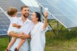Solar energy concept. A young, happy family is standing near solar panels and holding an electric light bulb.