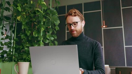A young red-haired guy, a programmer or entrepreneur in glasses, works in a stylish cafe behind a laptop. Freelancer works remotely. Online communication. Small business.