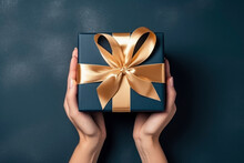 overhead view, womans hands holding a luxury gift box with gold large satin bow against a dark blue background. Close up. New Year present.
