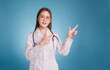 Young asian doctor wear medical uniform lab coat and stethoscope on blue background. Smiling female doctor pointing side to empty copy space for healthcare product or show banner rules safety.