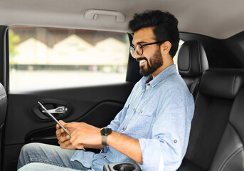 Wall Mural - Cheerful eastern guy using digital tablet while sitting in taxi