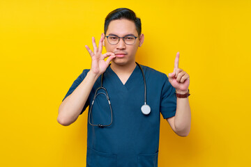 Wall Mural - Serious young Asian man doctor or nurse wearing a blue uniform and glasses makes a zipping mouth gesture, promising to keep secret, pointing up isolated on yellow background