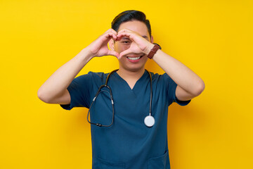 Wall Mural - Smiling professional young Asian man doctor or nurse wearing a blue uniform and stethoscope showing heart shape with hands isolated on yellow background. Healthcare medicine concept