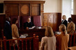 © AnnaStills - Group of witnesses, attorneys, lawyers, prosecutor and suspect standing in front of experienced blond female judge in mantle passing judgement