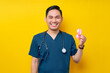 © Bangun Stock Photo - Smiling professional young Asian male doctor or nurse wearing a blue uniform and stethoscope holding a pink silk ribbon symbol isolated on yellow background. Breast Cancer Awareness concept