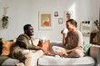 Side view portrait of young African American couple enjoying conversation sitting on comfortable couch in cozy home and drinking coffee
