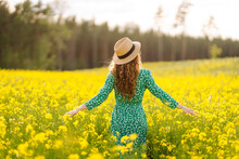 A Happy Woman In A Hat Walks Through A Blooming Rapeseed Field. Beautiful Woman Posing In A Rapeseed Field On A Sunny Day. Concept Of Nature, Relaxation.