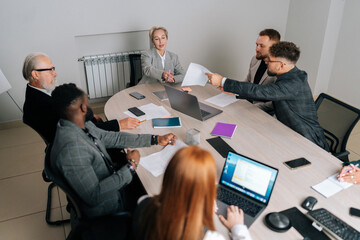 Top view of serious middle-aged female corporate leader giving papers report plan to diverse-aged team business people, discuss paperwork at group meeting. Businesswoman giving presentation report.