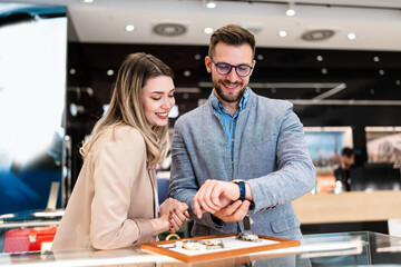 Elegant middle age businessman choosing and buying his new expensive watch. Beautiful young female seller helps him to make good decision. Fashion style and elegance concept.