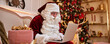 Santa Claus sitting at home and reading email on laptop with New Year wish list near Christmas tree