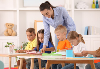 Sticker - Nursery teacher with group of cute little children drawing and cutting paper at desks in kindergarten. Playtime activities