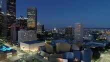 Aerial Backward Shot Of Walt Disney Concert Hall Amidst Modern Buildings In City Against Sky At Night - Los Angeles, California