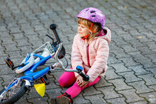 Cute Little Girl Sitting On The Ground After Falling Off Her Bike. Upset Crying Preschool Child With Safe Helmet Getting Hurt While Riding A Bicycle. Active Family Leisure With Kids.