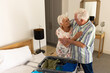 Happy caucasian senior couple packing suitcase and embracing in sunny bedroom at home