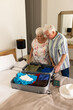 Happy caucasian senior couple packing suitcase and embracing in sunny bedroom at home