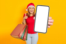 Smiling Young Asian Woman In Santa Claus Hat Holding Blank Screen Phone And Shopping Bag While Thumbs Up Isolated On Yellow Background
