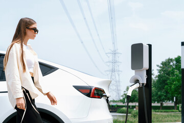 Young woman recharge EV car battery at charging station connected to power grid tower electrical industrial facility as electrical industry for eco friendly vehicle utilization. Expedient