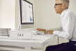 © Studio Romantic - White sign with the word TAX on the table on the background of a serious busy senior man who works sitting at a laptop and checks the register of taxpayers. Business and finance concept.