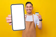© Bangun Stock Photo - Excited young Asian man 20s barista barman employee wearing brown apron working in coffee shop, holding credit bank card and showing smartphone blank screen isolated on yellow background