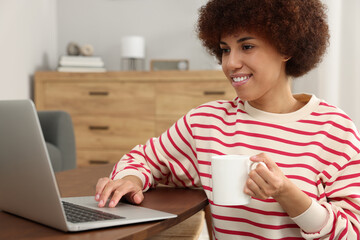 Wall Mural - Beautiful young woman using laptop and drinking coffee at wooden table in room