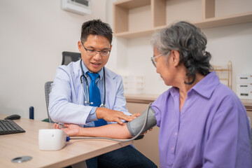 Asian caregiver doctor using digital pressure gauge measuring blood pressure of elderly woman patient. Young female nurse in uniform gives consultation during home visit. Medical and health concept.