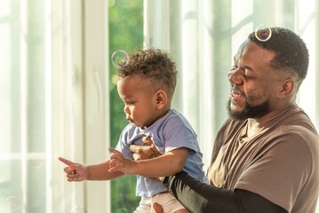 Wall Mural - Happy african black parents dad father throw baby son on piggyback and neck riding teasing fun near window. Black baby son and daddy enjoy teasing kiss cheek and neck riding at window light