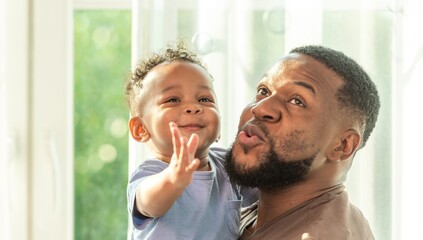 Wall Mural - Happy african black parents dad father throw baby son on piggyback and neck riding teasing fun near window. Black baby son and daddy enjoy teasing kiss cheek and neck riding at window light