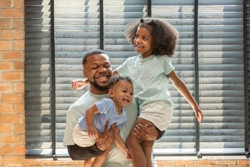 Wall Mural - Happy african black dad father carry hold baby son and daughter on arm and piggyback or neck riding teasing fun near window. Black baby son and daughter enjoy teasing daddy at window light