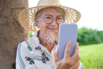 Wall Mural - Closeup portrait of smiling attractive senior woman with hat and eyeglasses sitting in public park using mobile phone - caucasian lady enjoying free time holding cellphone