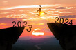Greetings and have a great Christmas and new year in 2024.silhouette of a man leaping from a cliff in 2023 to one in 2024 under a cloudy and sunny sky.