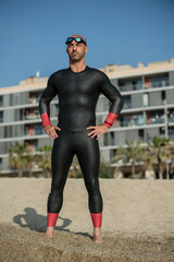 Wall Mural - Confident man wearing a wetsuit standing in the shore ready for swimming. Professional athlete staring at the ocean in the beach with urban background.