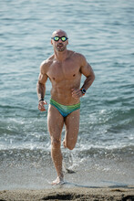 Sexy Muscular Man In Colorful Speedo Running Out Of The Sea Looking At Camera. Professional Male Athlete Getting Out Of The Water Wearing Swim Glasses At The Beach.