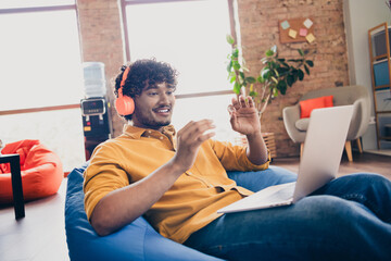 Poster - Photo portrait of attractive young man wear yellow shirt sit blue bean bag talk video call headphones modern workplace room home design