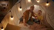 © kinomaster - Playful father teaches his son to howl like wolves. A man and a boy sit in a garland lit tent in a dark living room close up. A father and son's evening together. Concept of happy childhood.