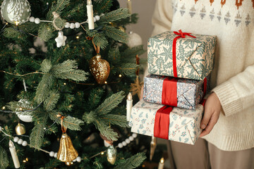 Wall Mural - Woman in sweater holding stylish christmas gifts with red ribbon at modern decorated vintage tree in scandinavian room. Merry Christmas and Happy holidays!