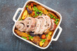Rolled sliced pork roasted in white casserole dish with potatoes, vegetables and herbs on blue dark concrete rustic background top view. Baked pork roll with vegetables for dinner.