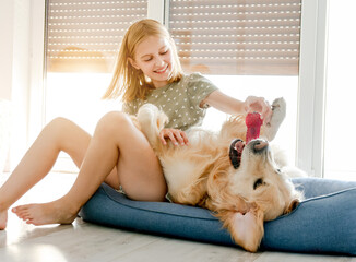 Wall Mural - Cute preteen child girl with golden retriever dog playing at pillow at floor together. Pretty female kid with doggy pet labrador and toy at home