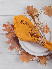 Wall Mural - Autumn table setting with decorations on white wooden background. Empty plate, cutlery, yellow linen napkin and dry leaves. Autumn holiday mood, Halloween, Thanksgiving concept. Top view