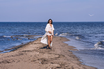 Wall Mural - Portrait of attractive brunette girl with long hair posing on a deserted beach. She wears white shorts, a top, a long white cape. She is looking to the camera.