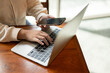© bongkarn - Close-up image of a woman using her smartphone while working on her laptop at a table in a cafe.