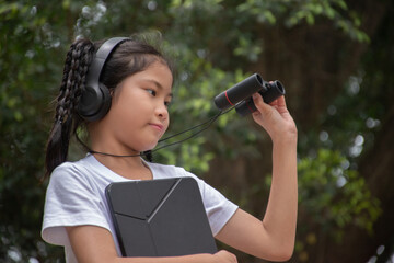 Asian cute girl holds tablet in chest and mini binoculars to watch birds, insects, leaves and small things in the park, recreational activity and nature study concept.