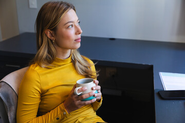 Caucasian woman sitting at desk, working from home with laptop and drinking coffee in sunny room