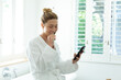 Happy caucasian woman in bathrobe brushing teeth and using smartphone in sunny bathroom