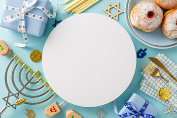  Hanukkah dining table concept. Overhead shot featuring sufganiyot, napkin, cutlery, Star of David, gift boxes, menorah, gelt, dreidel on a soft blue surface with circular card for text or promotion